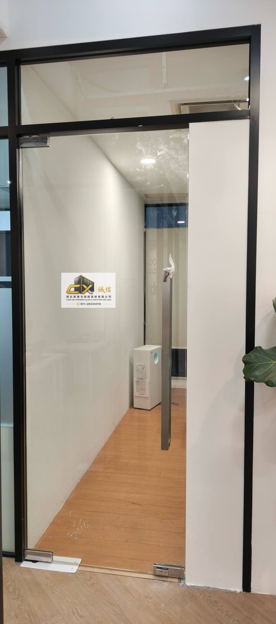 A black-framed glass door and side panel opening onto a corridor with wood-effect flooring.
