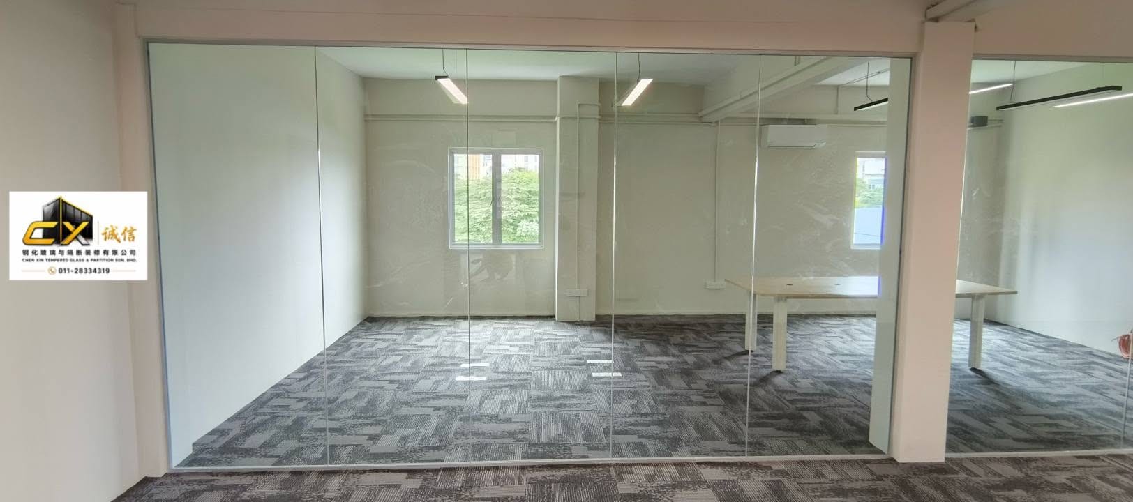 A frameless tempered-glass partition enclosing an empty carpet-tiled office, with daylight from two windows.