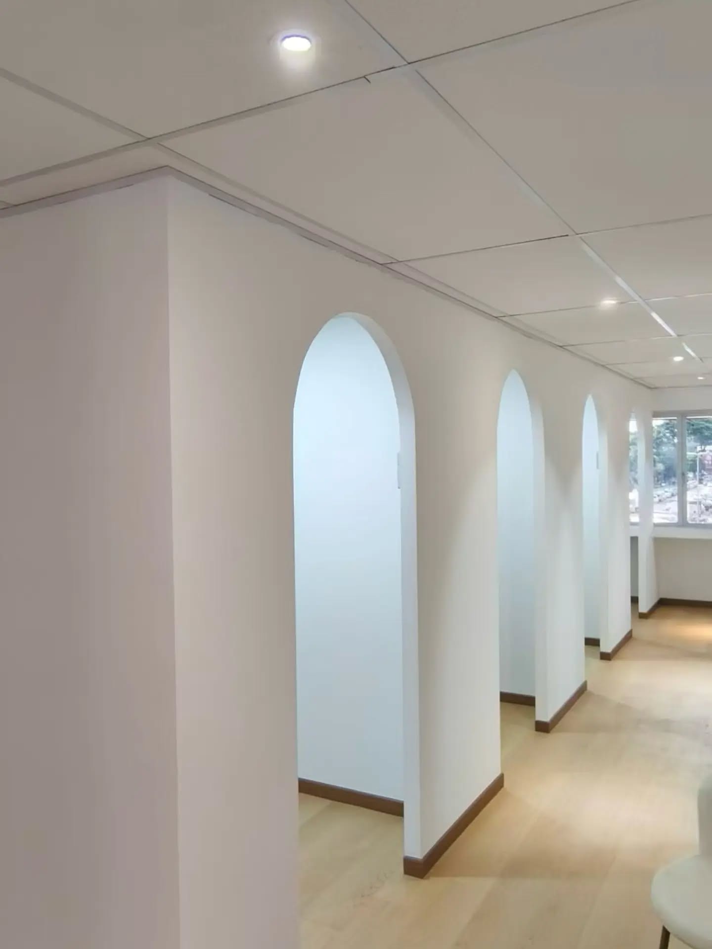 An office corridor formed by gypsum-board partitions with arched openings, wood-effect flooring and recessed downlights.