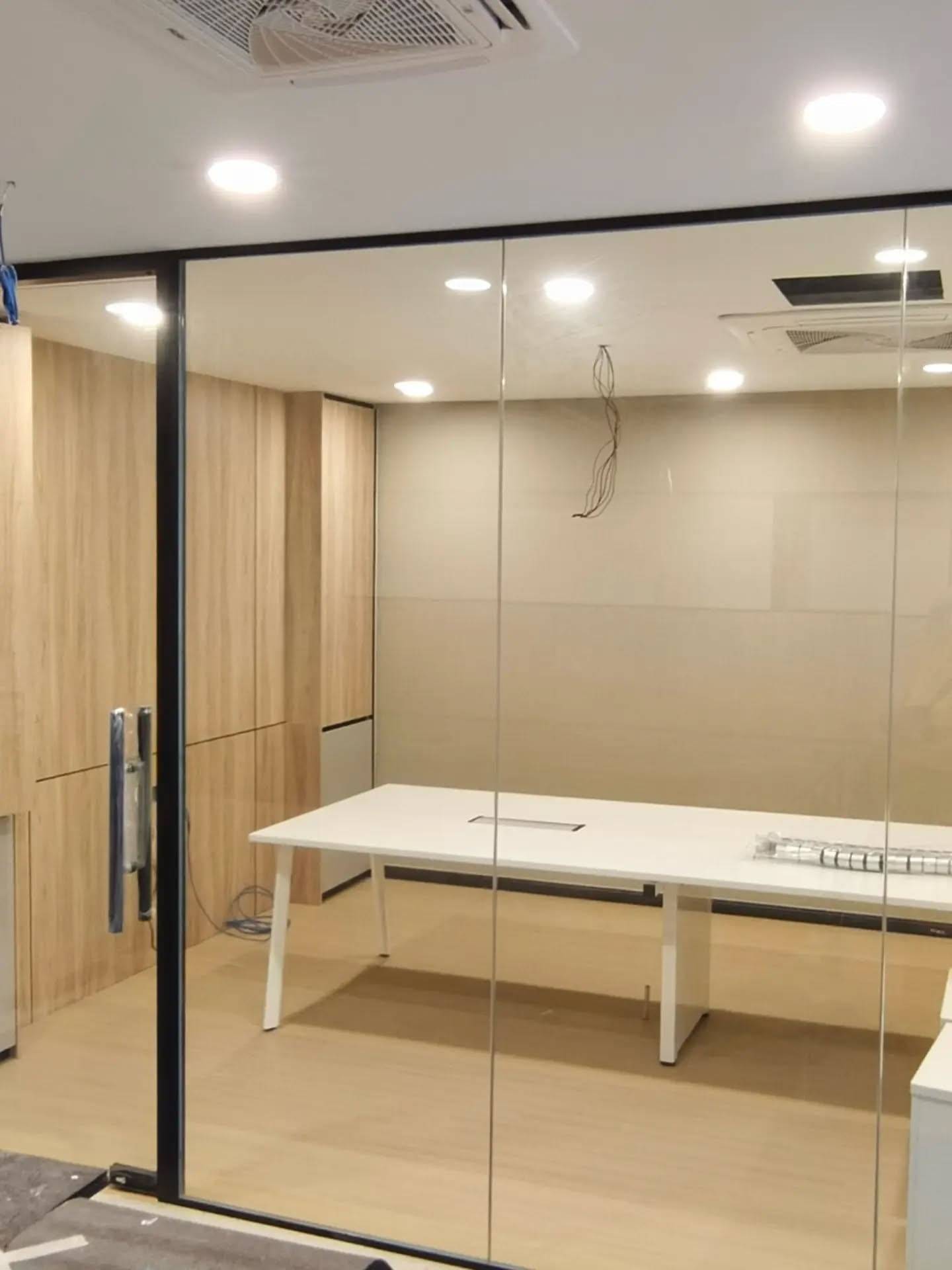 A meeting room enclosed in black-framed tempered glass, with a long table inside and wood-effect flooring.