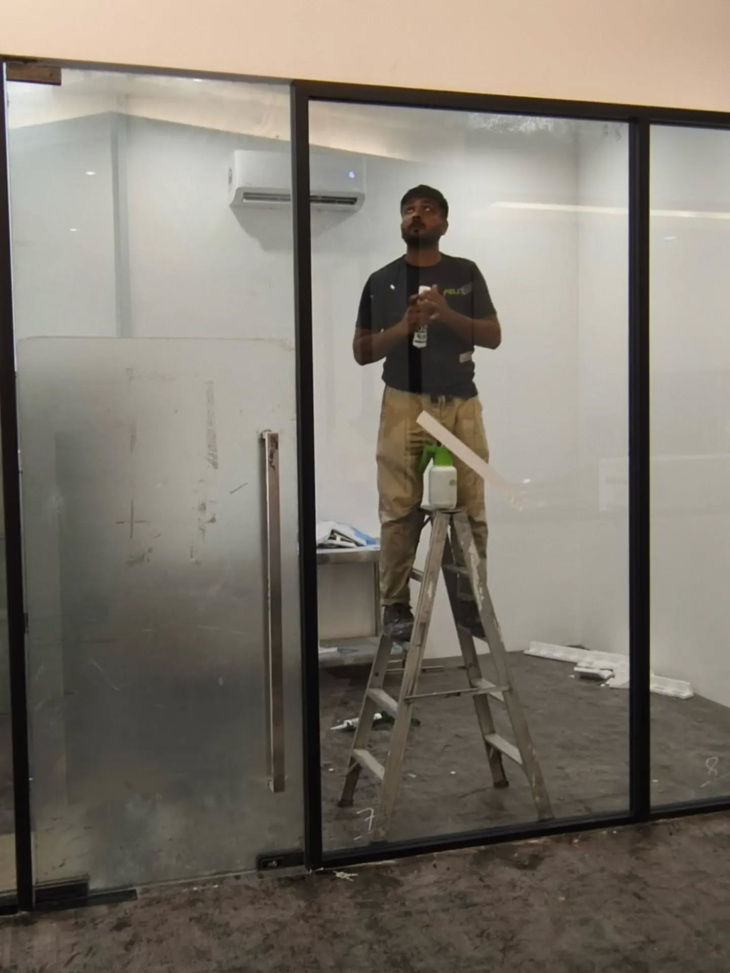 An installer on a step-ladder behind a black-framed glass door leaf that is already hung.
