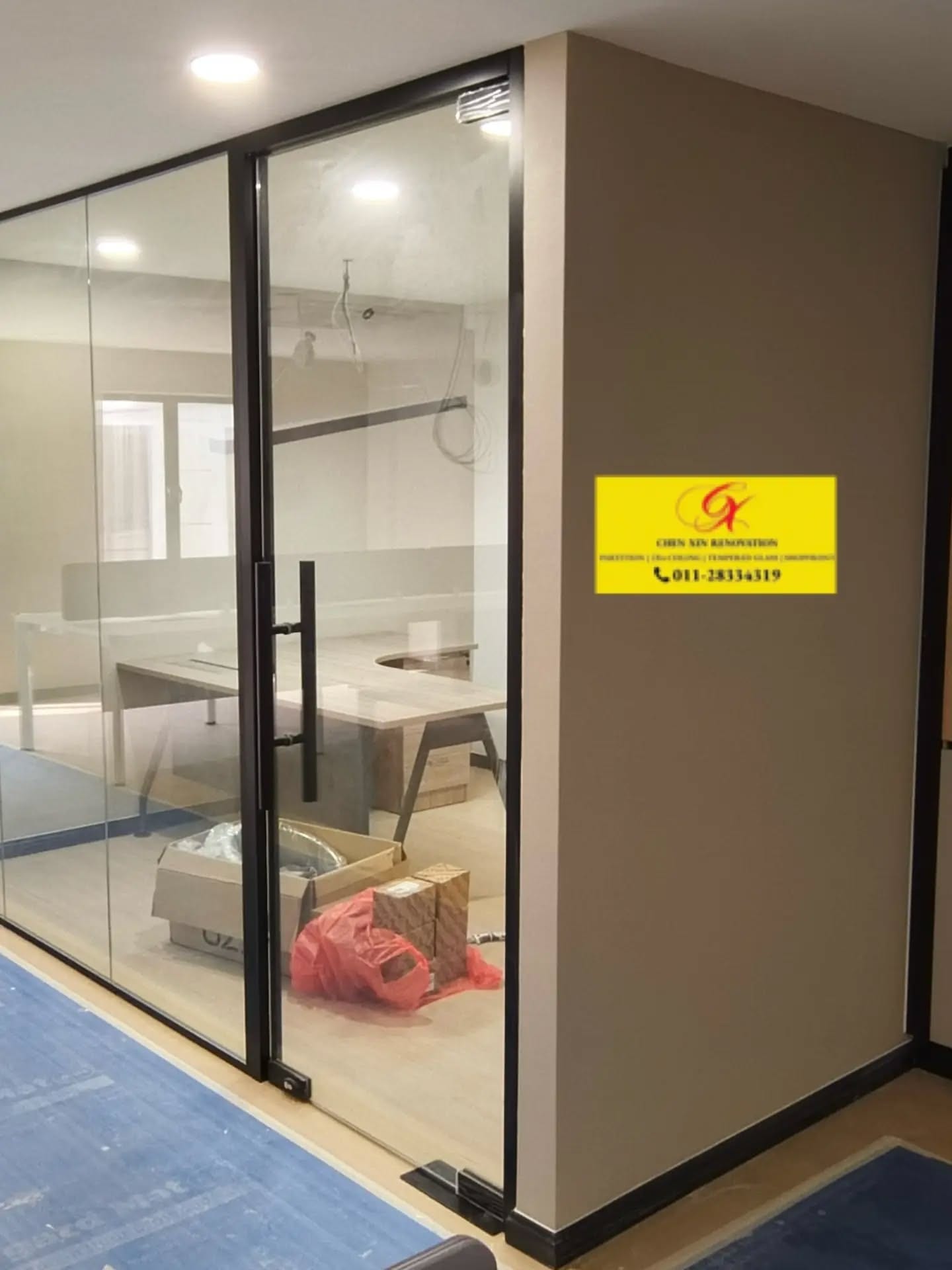 A black-framed glass partition standing against a white wall, with blue protective sheeting over the floor.