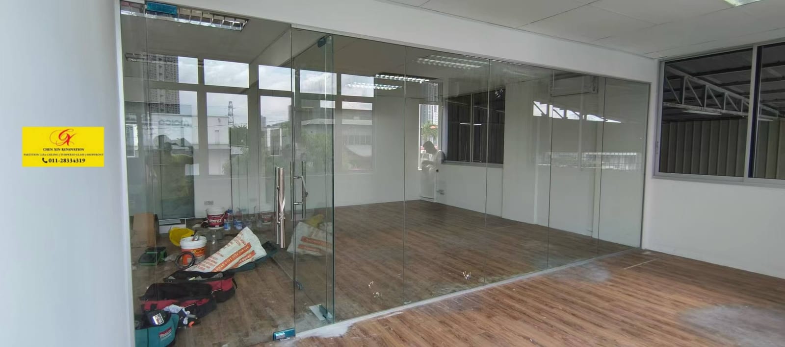 The same office mid-installation: the frameless glass run already set on its floor fixings, tool bags and cement bags still on the protected floor, the ceiling not yet closed.