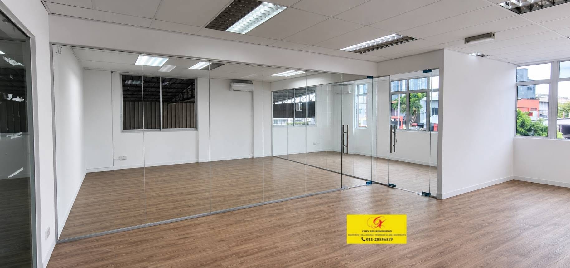 A finished office divided by a run of frameless tempered-glass partitions with a glass door, wood-effect flooring and a suspended ceiling.